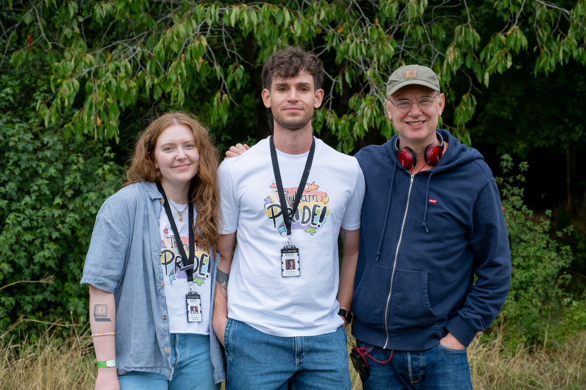 Three people smiling outdoors in front of trees, two wearing "Pride" shirts and lanyards, one with headphones and a cap, suggesting a casual event or festival in a natural setting.