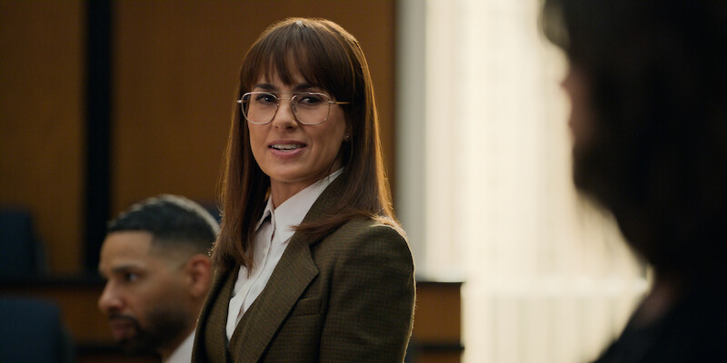 A woman in professional attire stands in a courtroom, speaking with confidence. Two people are seated nearby, contributing to a serious, focused atmosphere. The lighting is natural, highlighting a formal and intense legal setting.