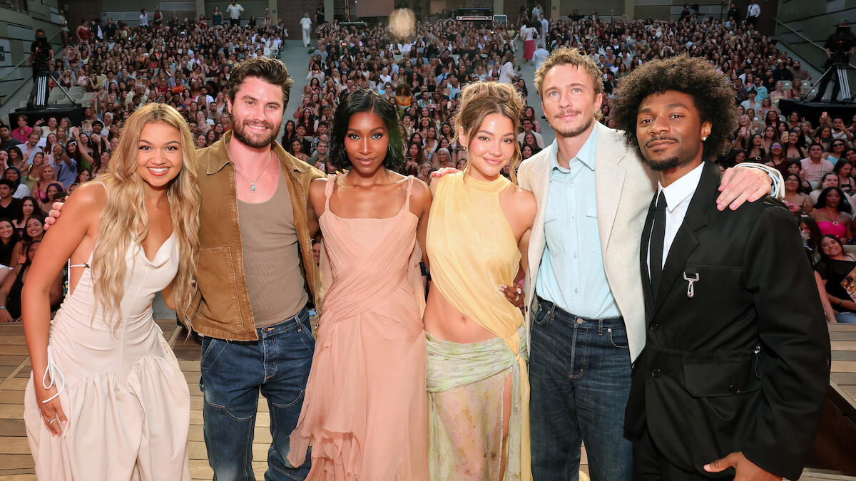 Six people pose together at the center of a stage, standing arm-in-arm and smiling. Behind them is a large audience seated in an amphitheater setting under daylight conditions.