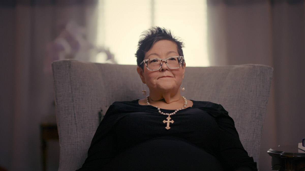 Woman with short dark hair and glasses sits in a gray armchair, wearing a black top and a large cross necklace, with a window and soft curtains in the background of a warmly lit room.