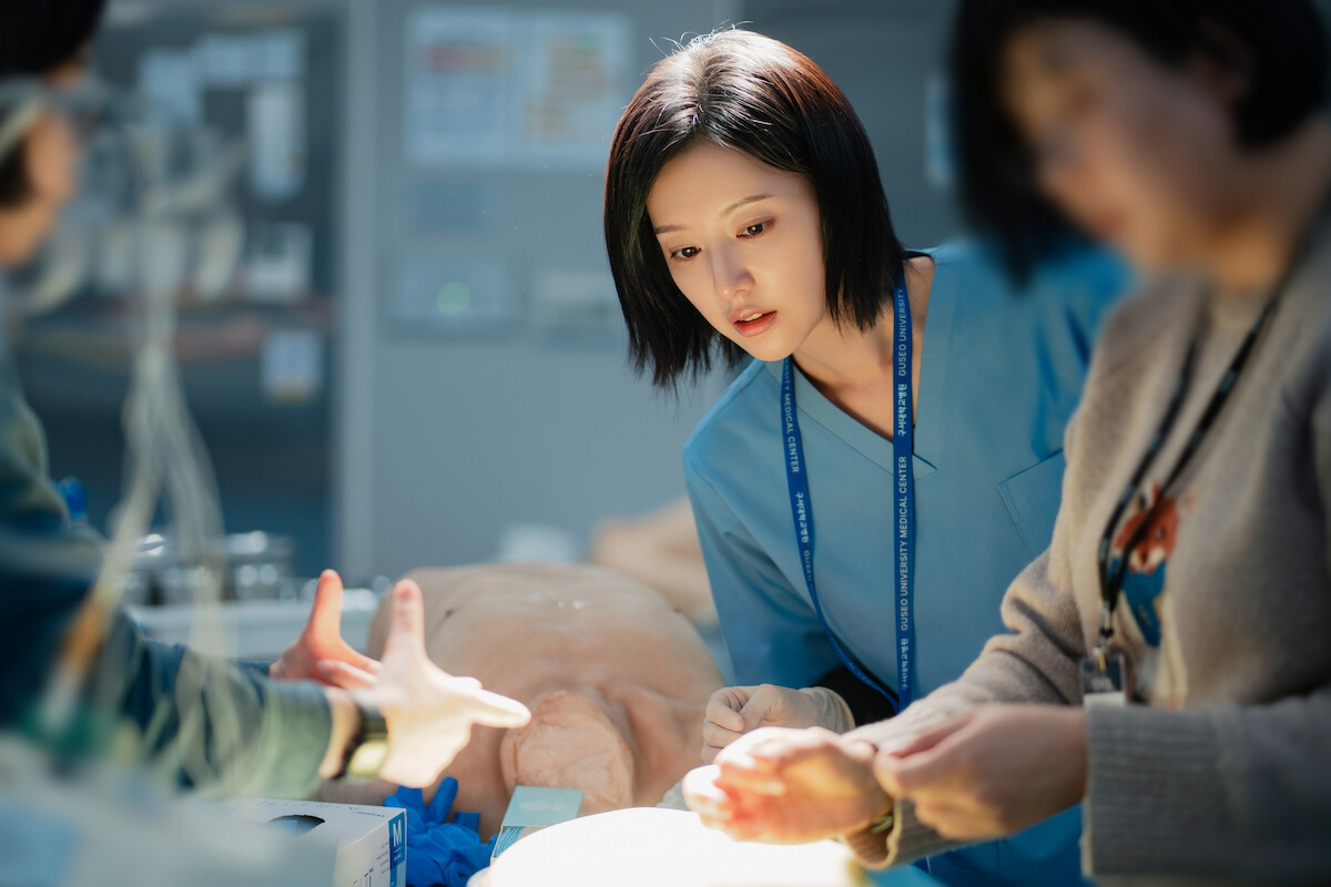 Uma mulher sul-coreana de cabelos pretos e curtos veste roupas médicas e observa uma operação