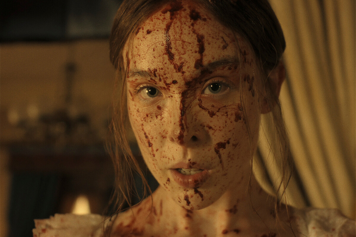 A woman with blood splattered on her face stands indoors, looking directly at the camera, with vintage curtains and warm lighting in the background.