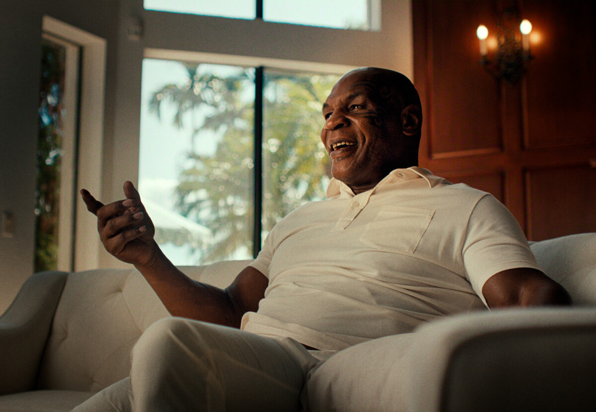 O lutador Mike Tyson, um homem negro e de cabelos raspados, veste camisa branca e gesticula para a câmera sentado em um sofá de sua casa