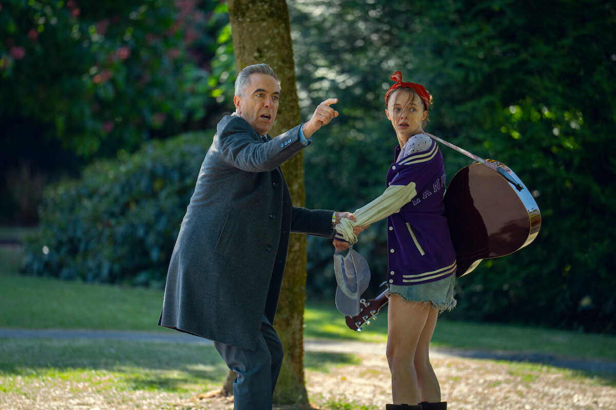 A man in a suit and a woman with a guitar and casual clothes stand in a park, holding hands and looking surprised or alarmed, surrounded by trees and greenery.
