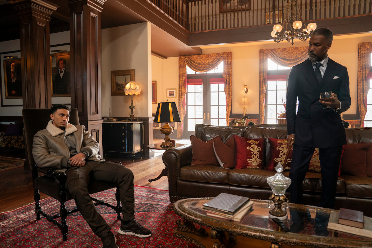 Two men in a luxurious living room; one is seated casually in a chair while the other stands by a sofa holding a drink. The room has elegant décor, large windows, wooden columns, ornate furniture, warm lighting, and a red rug.
