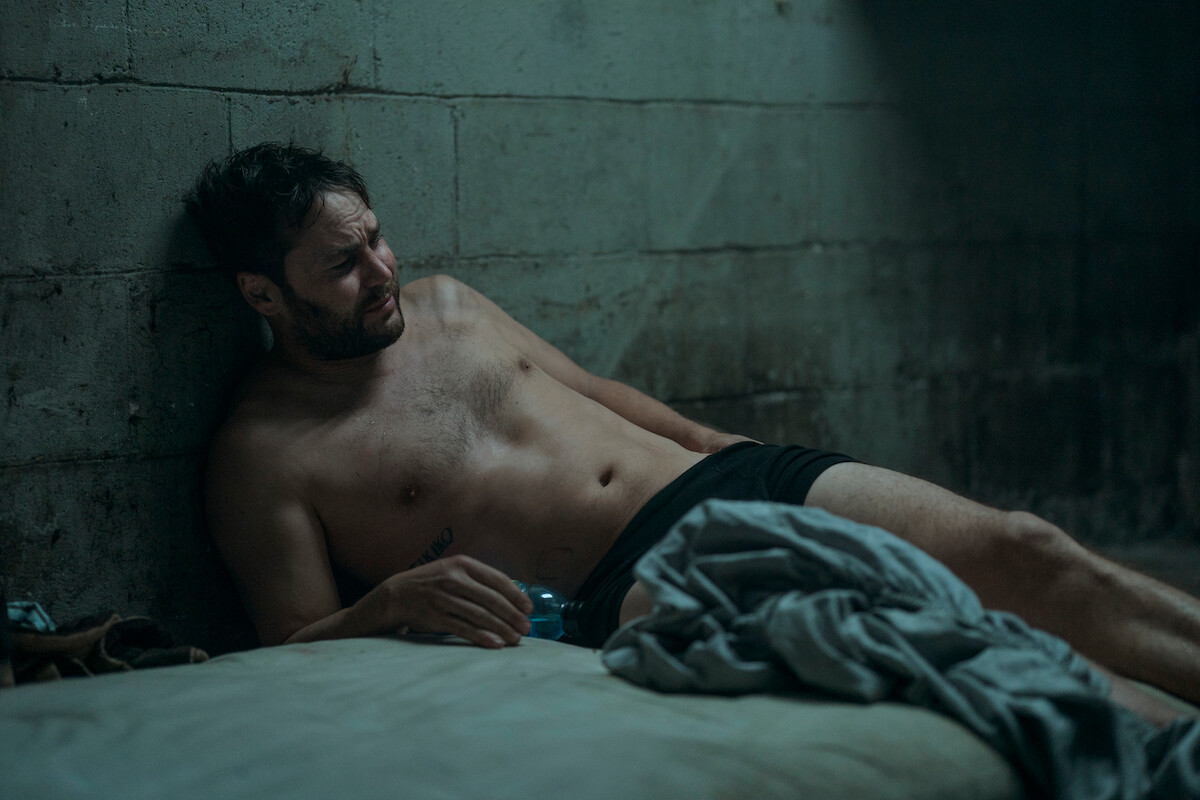 Taylor Kitsch as Glen Kryger lays down on a gray mattress with a pained expression in Season 1 of “Painkiller.’