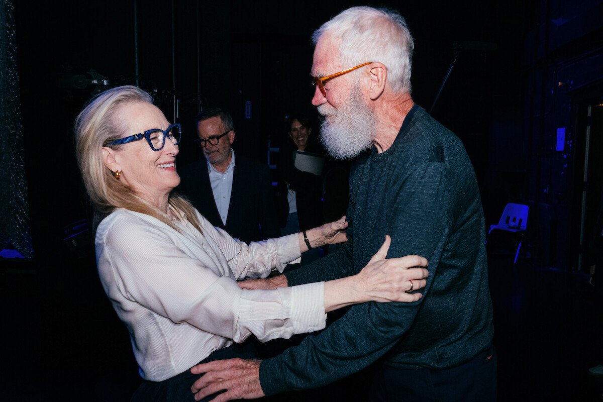Meryl Streep and David Letterman smiling and embracing each other warmly backstage in a dimly lit setting, with a few other people and stage equipment visible in the background.
