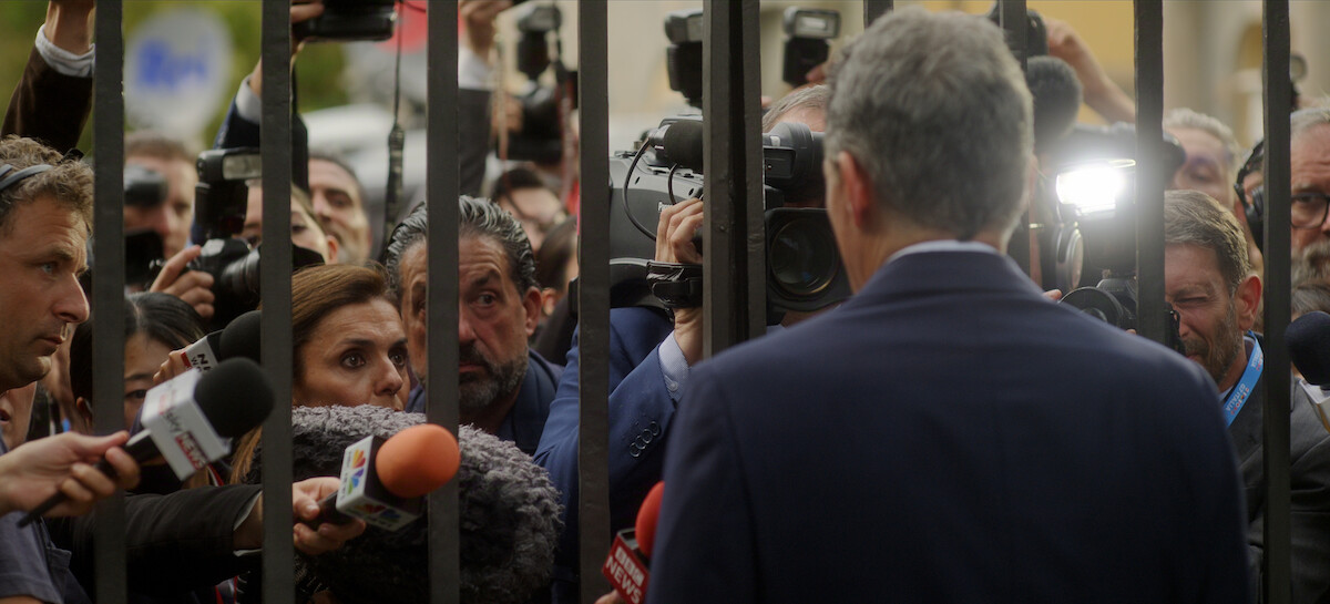 A man in a suit speaks to a crowd of journalists and photographers behind a metal fence outdoors, with microphones and cameras pointed toward him.