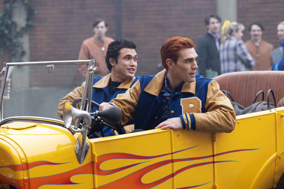 Charles Melton as Reggie Mantle and KJ Apa as Archie Andrews sit in a vintage yellow car painted with red flames in Season 7 of ‘Riverdale.’