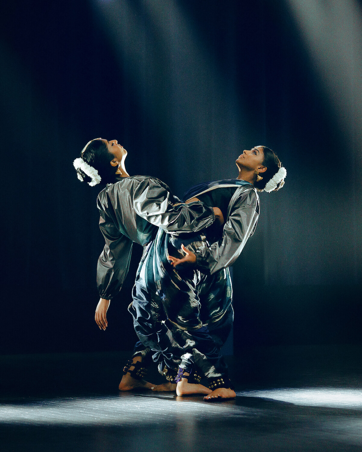 Two dancers in traditional attire perform an expressive pose on a dimly lit stage, with dramatic spotlights highlighting their coordinated movement and elegant costumes against a dark background.