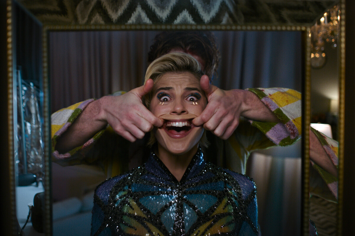 Person in sequined outfit smiling in a mirror as another person, partially visible, stands behind them stretching their mouth into a grin; indoor setting with curtains, soft lighting, and patterned decor in the background.