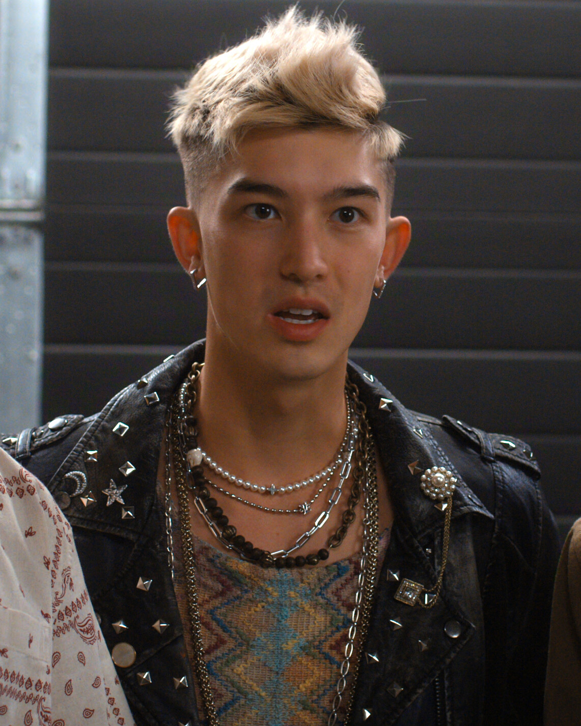 Young person with short, bleached hair, wearing multiple necklaces, earrings, and a studded leather jacket, is indoors in front of a dark industrial background, looking surprised or engaged.