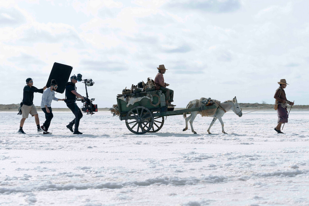 A film crew follows a donkey pulling a cart with a seated man across a vast, white, barren landscape under a cloudy sky, capturing a cinematic scene in progress.