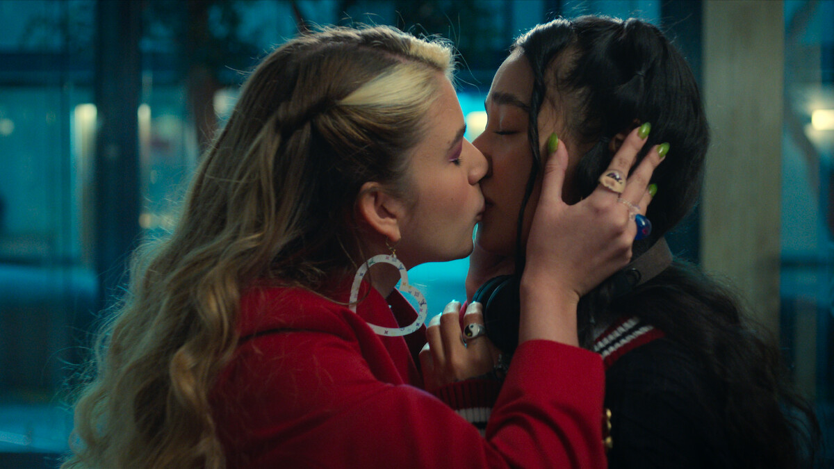 Two young women with styled hair and colorful outfits share an affectionate kiss indoors at night, emotional moment with city lights softly glowing in the background through large windows.