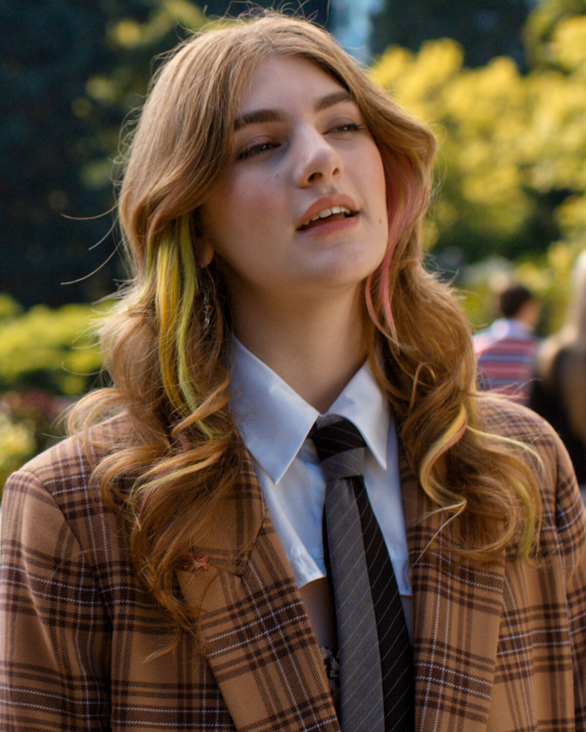 Person with wavy hair and colored streaks wearing a plaid jacket, white shirt, and tie, standing outdoors in a park-like setting with greenery and blurred people in the background.
