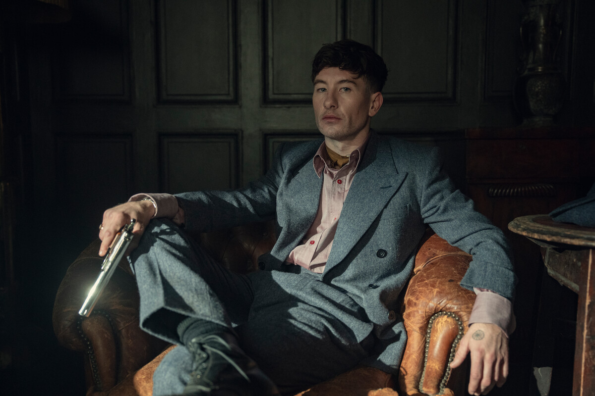 Man in a gray suit sitting in a vintage leather armchair, holding a gun, in a dimly lit room with dark paneled walls, creating a tense and dramatic atmosphere.