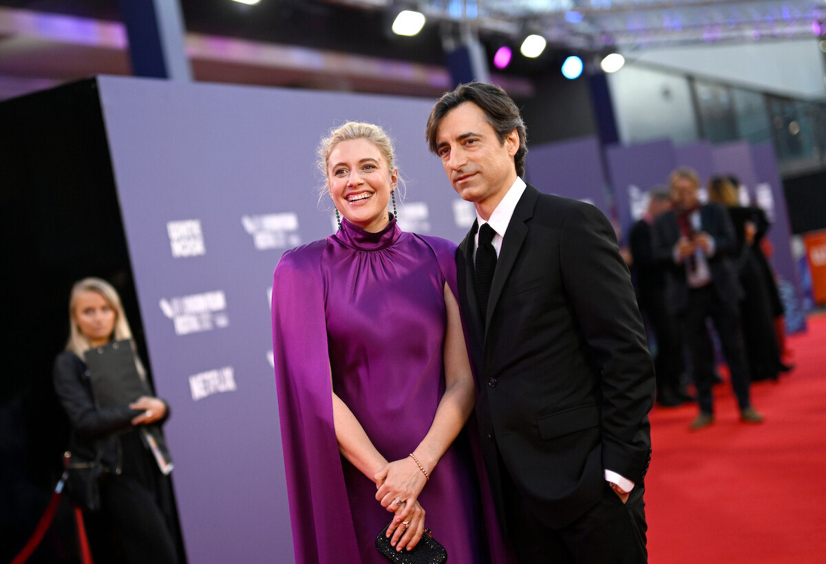Greta Gerwig, in a purple dress, and Noah Baumbach, in a black suit, stand on a red carpet.