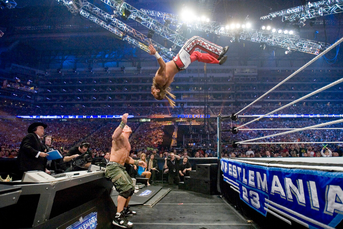 A wrestler performs a high-flying move off the ropes toward an opponent outside the ring during WrestleMania 23, with a large energetic crowd and bright arena lights creating an intense, action-packed atmosphere.