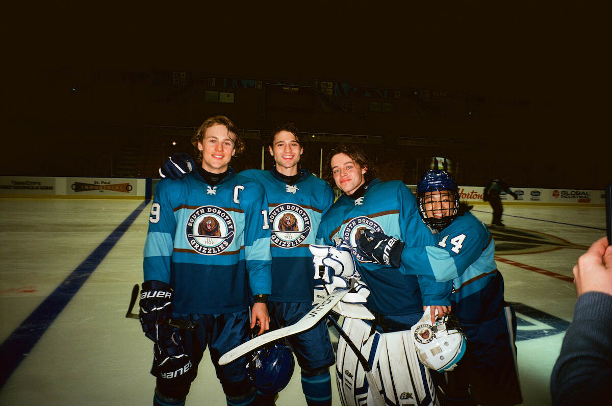 Four hockey players in blue uniforms posing together on an ice rink, smiling for a photo. The rink is indoors with boards and advertisements visible in the background, suggesting a team game or practice session.