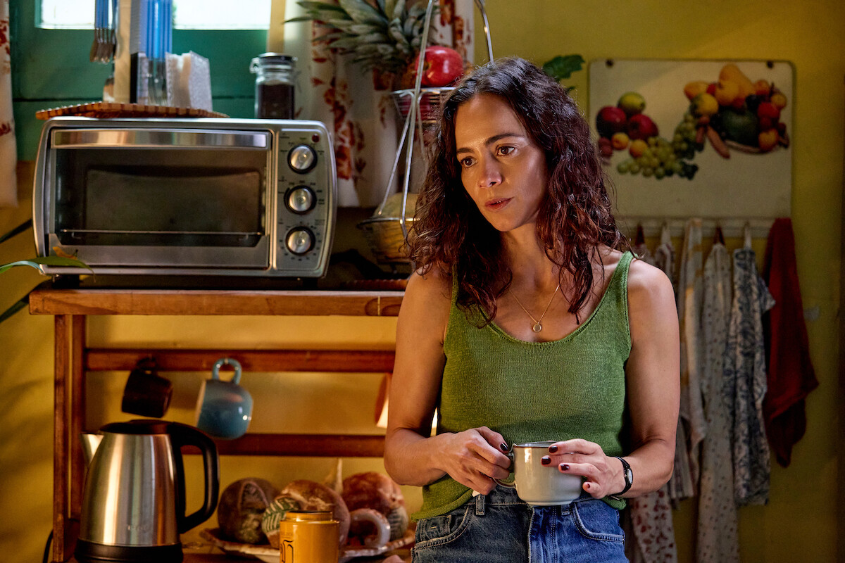 A woman in a green tank top stands in a cozy kitchen holding a mug. She is next to a toaster oven, kettle, bread, fruit decor, and hanging cups, with sunlight coming through a window behind her. The kitchen feels lived-in and homey.