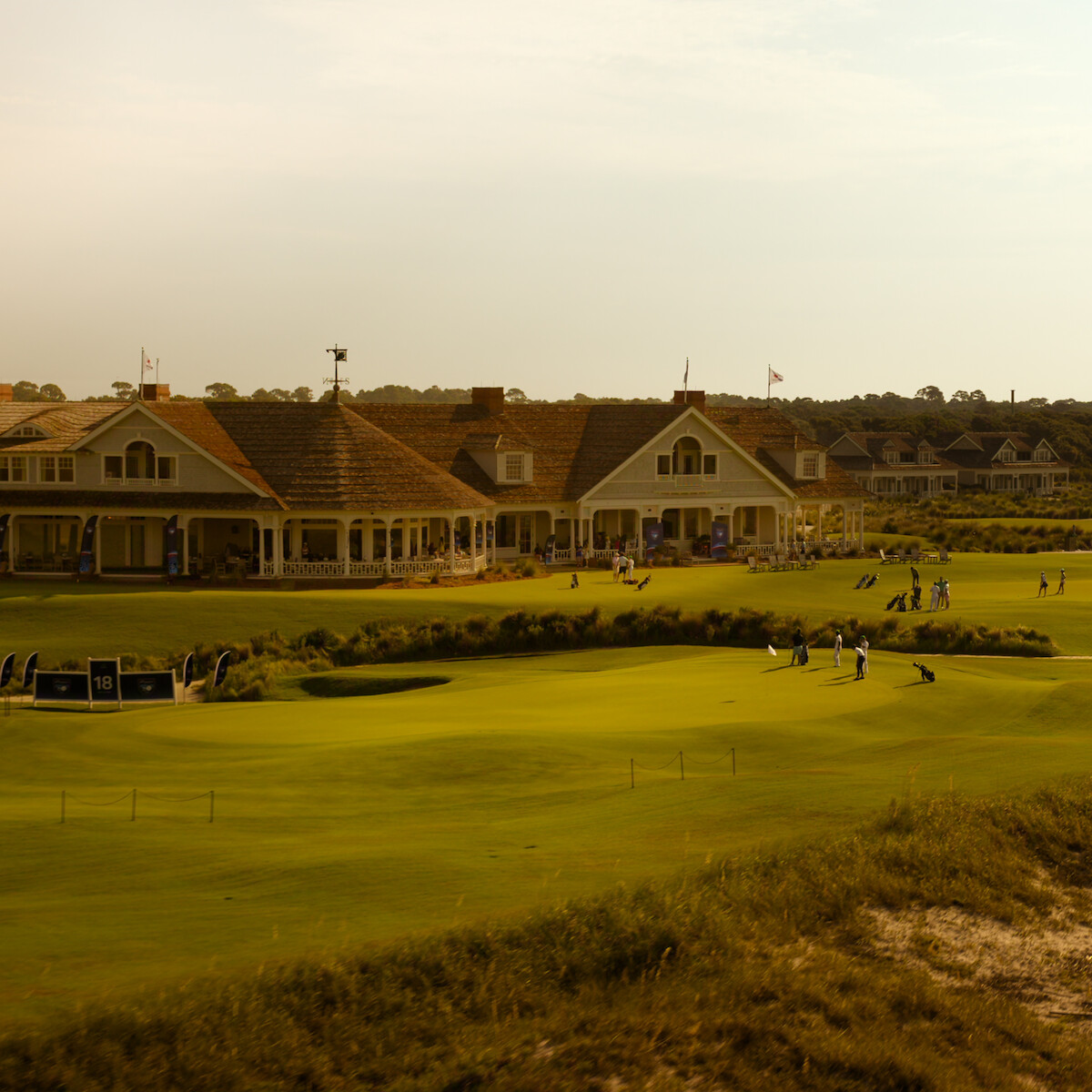 Golfers on a green near the 18th hole with golf bags, in front of a large clubhouse and cottages at sunset, surrounded by manicured lawns and landscaping.
