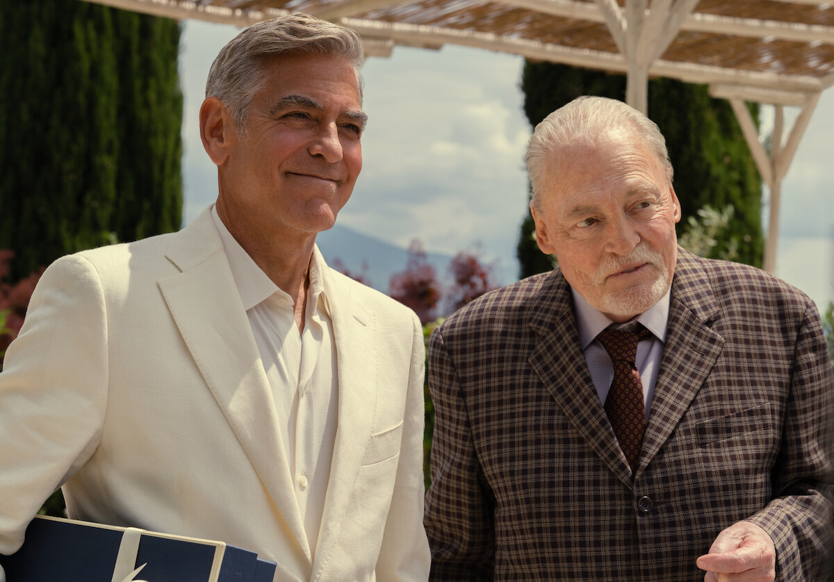 Two well-dressed older men stand outdoors under a pergola on a sunny day, surrounded by greenery and flowers, one smiling and the other looking thoughtful, creating a warm and relaxed atmosphere.