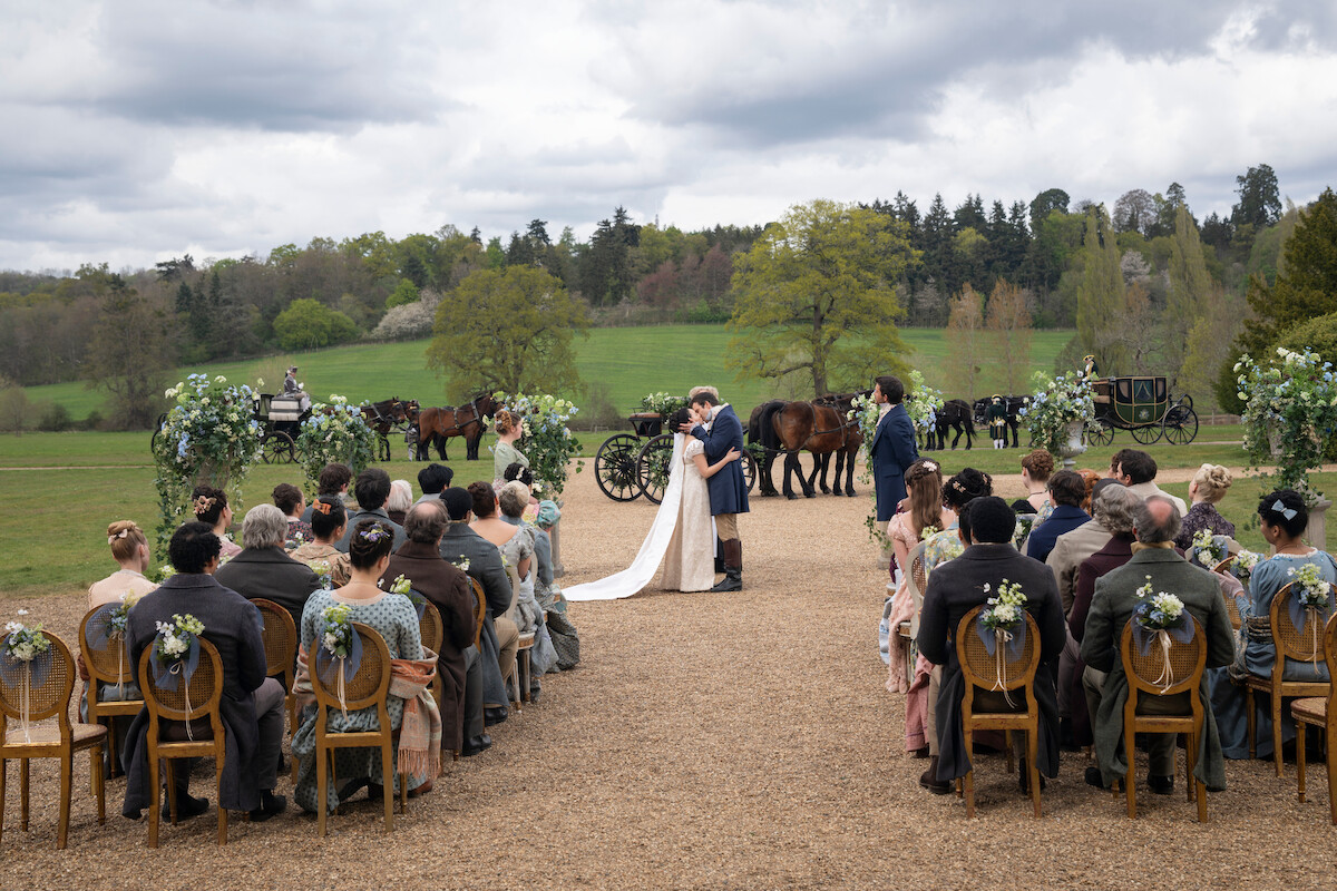 A bride and groom kiss at an outdoor wedding ceremony, surrounded by guests seated in decorated chairs. The setting is a green field with trees, carriages, and horses visible in the background under a cloudy sky.