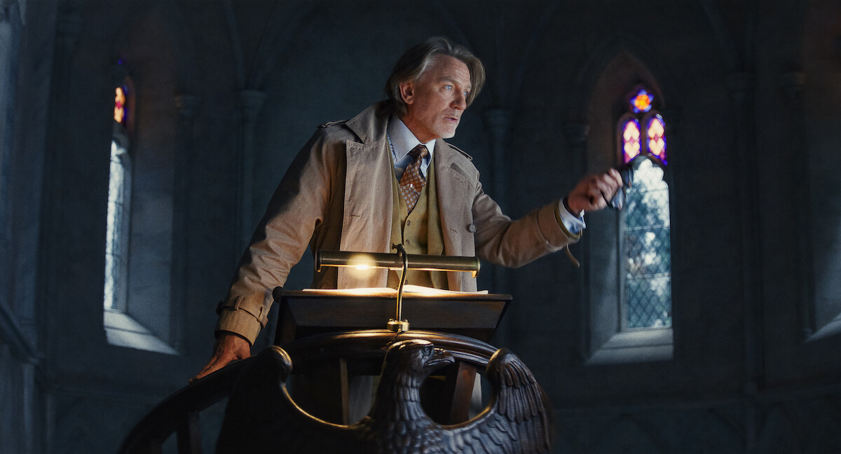 An older man in formal attire stands at a lectern shaped like an eagle in a dimly lit, gothic-style room with stained glass windows, creating a serious and dramatic atmosphere.