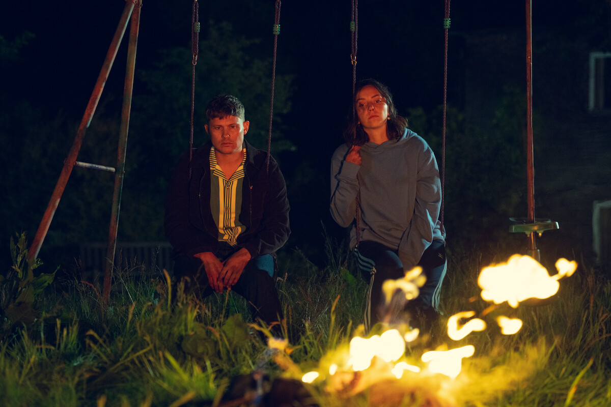 Two people sit on swings at night in a grassy outdoor area, faced with a campfire burning brightly in the foreground, with a dark background and some trees and buildings barely visible.