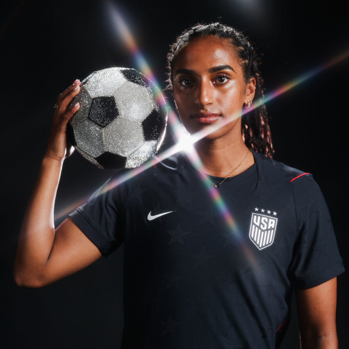 Female soccer player in dark USA uniform poses confidently in a studio holding a sparkling soccer ball, with dramatic lens flares and a dark background enhancing the athletic and artistic vibe.