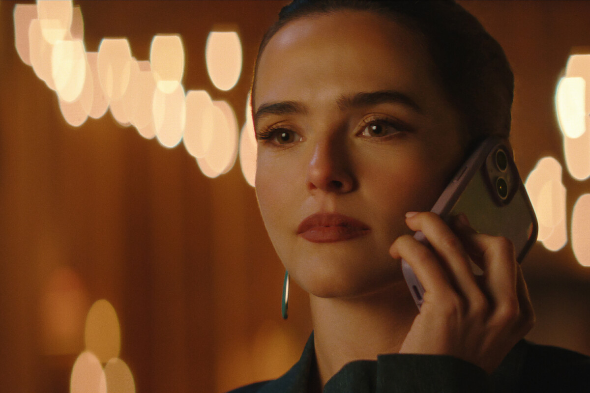 A woman with a serious expression holds a cellphone to her ear, speaking on a call. The background is softly lit with warm, blurred decorative lights, creating a cozy and intimate indoor atmosphere.