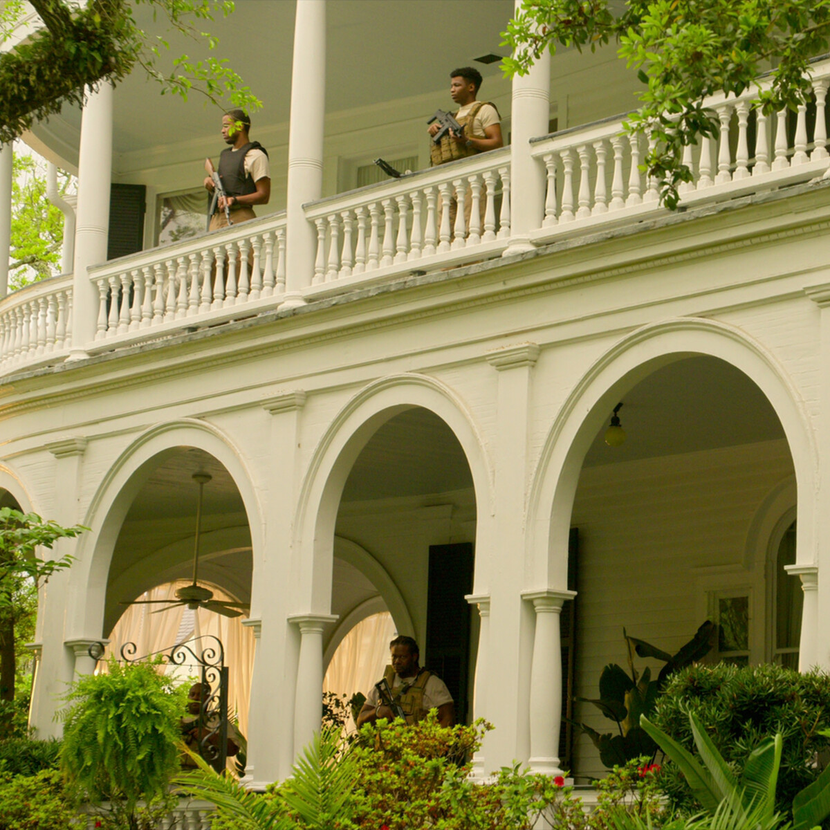 Three people with tactical gear stand on the porch and balcony of a large white house with arches and columns, surrounded by lush greenery in a daytime outdoor setting.