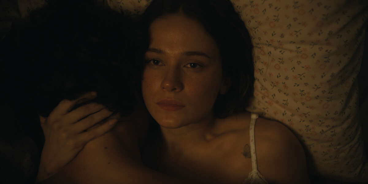 Woman lying in bed with a pensive expression, embracing another person whose face is not visible, dimly lit with floral bedsheets in the background.
