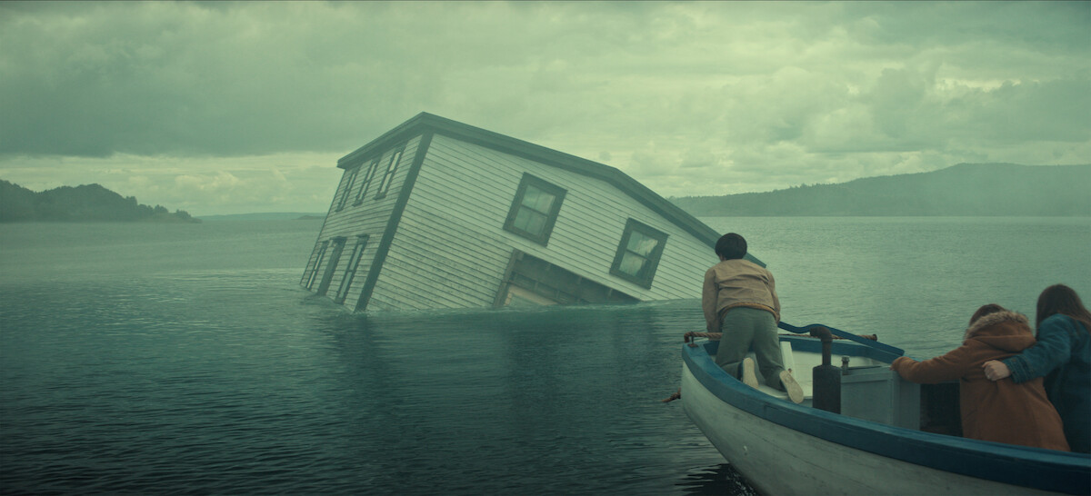 A partially submerged house tilts in a misty lake as three people in a small boat observe, with cloudy skies and distant hills creating a dramatic, mysterious atmosphere.