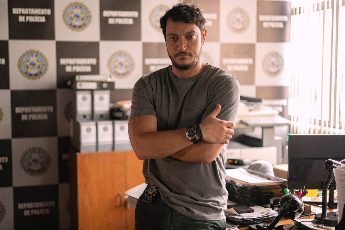 Um policial está em pé em frente a uma mesa de sua delegacia, vestindo camiseta cinza e calça comprida. Ao fundo é possível ver um computador e diversos objetos na mesa