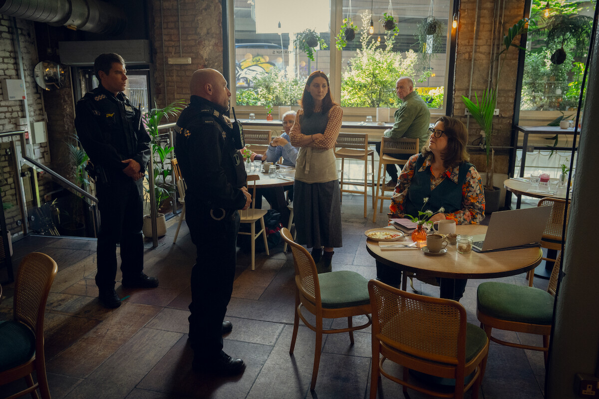 Two police officers speak with several people inside a cozy, plant-filled café with large windows, wooden chairs, tables, and natural light during the day.