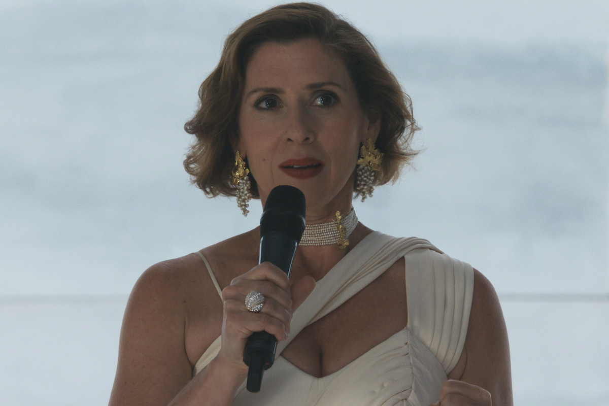 Woman in elegant white dress with gold earrings and necklace holding a microphone, speaking indoors against a softly lit background.