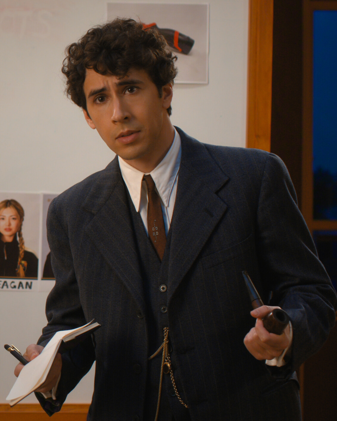 Man in a suit holding a notepad and marker, standing in front of a whiteboard with photos and notes, in an indoor setting with window and wooden frame in the background.