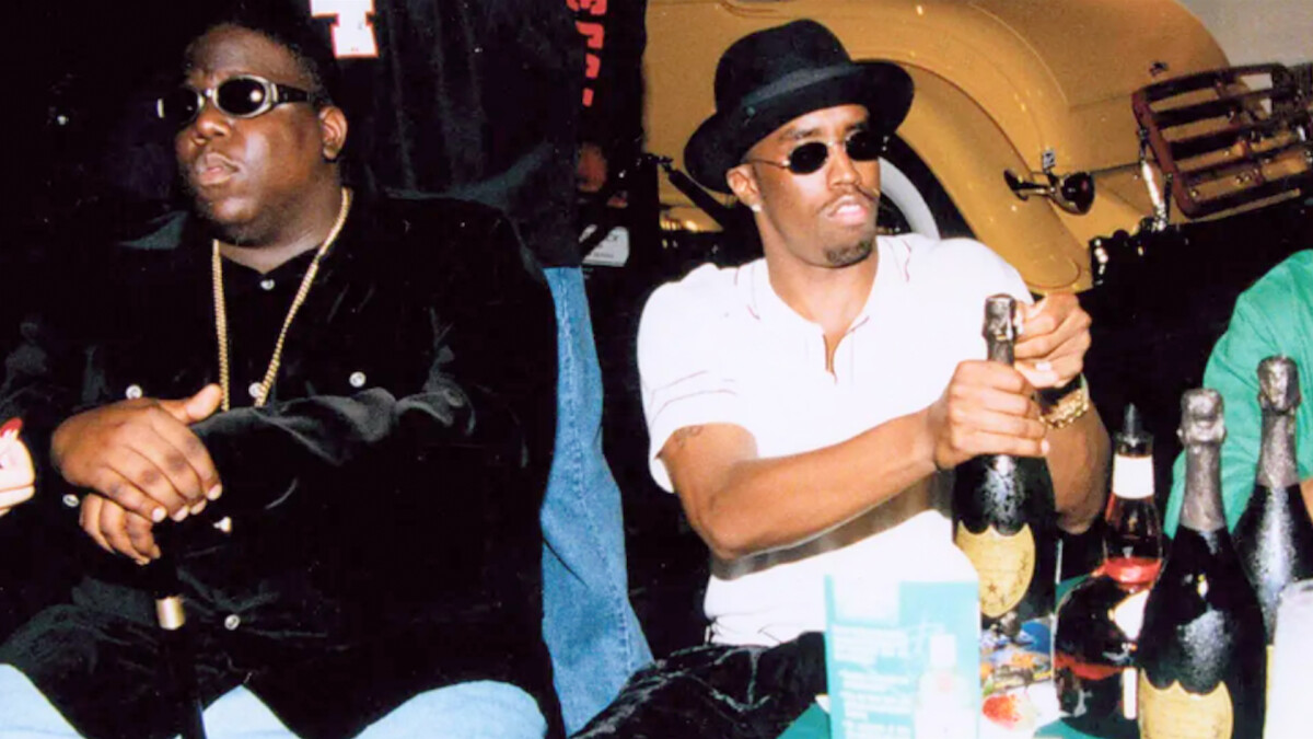 Two men in stylish outfits sit at a table with champagne bottles, one opening a bottle, in a lively, celebratory indoor setting with a vintage car in the background. The mood is festive and relaxed.