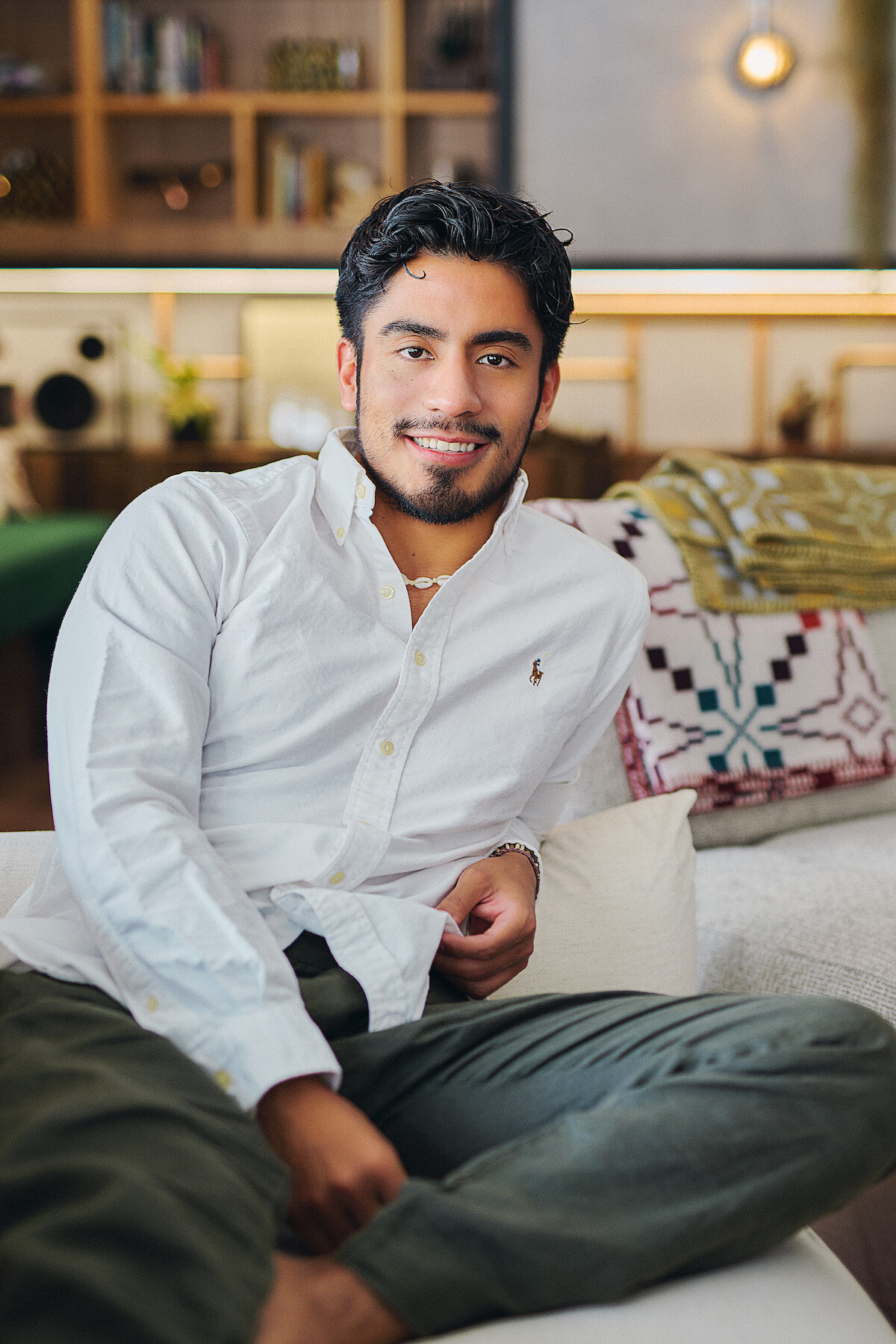 A young man in a white shirt sits relaxed on a modern sofa in a cozy, well-lit living room with shelves, decor, and patterned blankets, creating a warm and inviting mood.