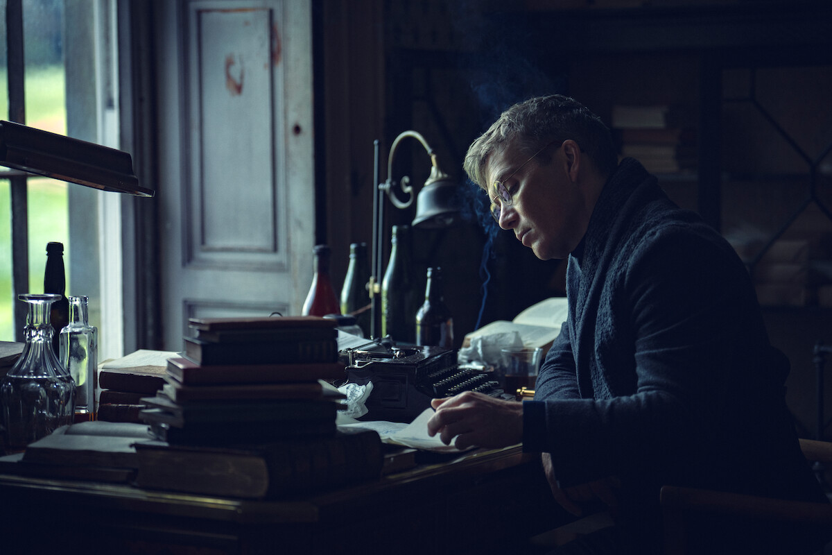 Man writing at a cluttered desk by a window in a dimly lit study, surrounded by stacks of books, bottles, a typewriter, and old-fashioned decor, creating a contemplative and vintage atmosphere.