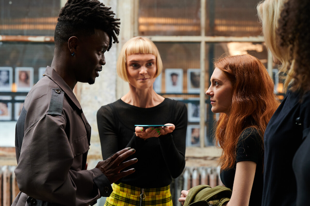 Emmanuel Imani as Wilbur Evans, Hebe Beardsall as Betty, and Emily Carey as Harriet Manners stand in a modeling agency facing one another in an image from the series ‘Geek Girl.’