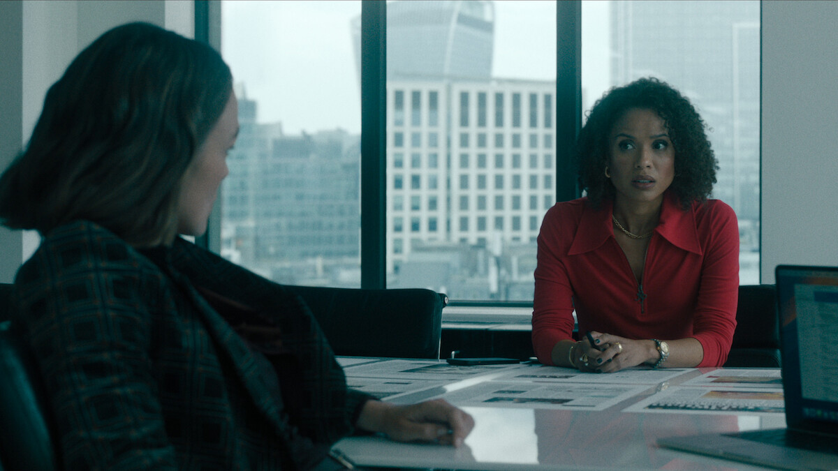 Gugu Mbatha-Raw wears a red blouse behind a desk while making a concerned expression opposite Kiera Knightley.