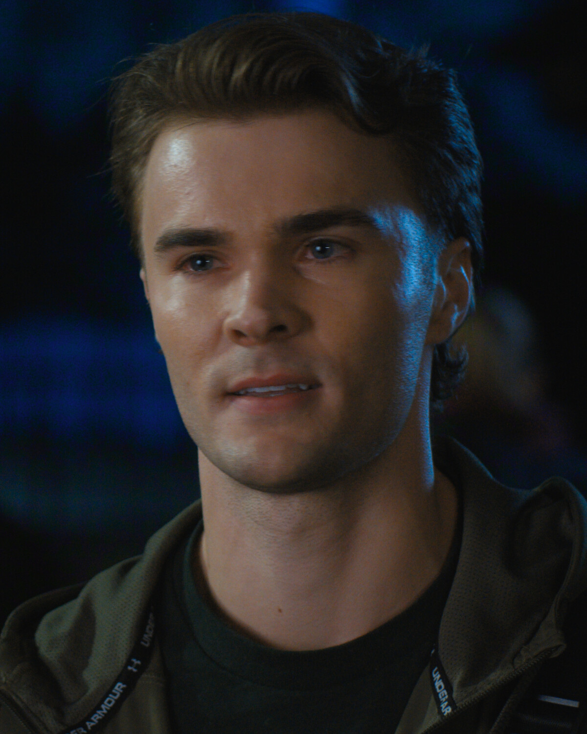 A young man with short brown hair stands outdoors at night, wearing a green jacket and dark shirt. The background is blurred with blue and black tones, suggesting an urban or nighttime setting.