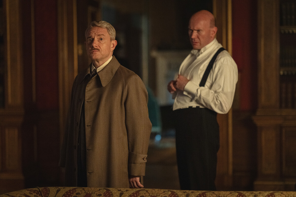 Two men stand in an elegant, dimly lit room with wood paneling and bookshelves; one wears a long coat and looks serious while the other adjusts his suspenders, adding a sense of tension to the scene.