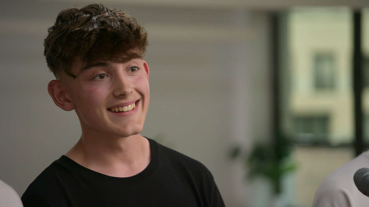 Smiling young man with brown hair wearing a black shirt, standing indoors in a bright room with blurred background, natural light, plants, and window. The mood is cheerful and relaxed.