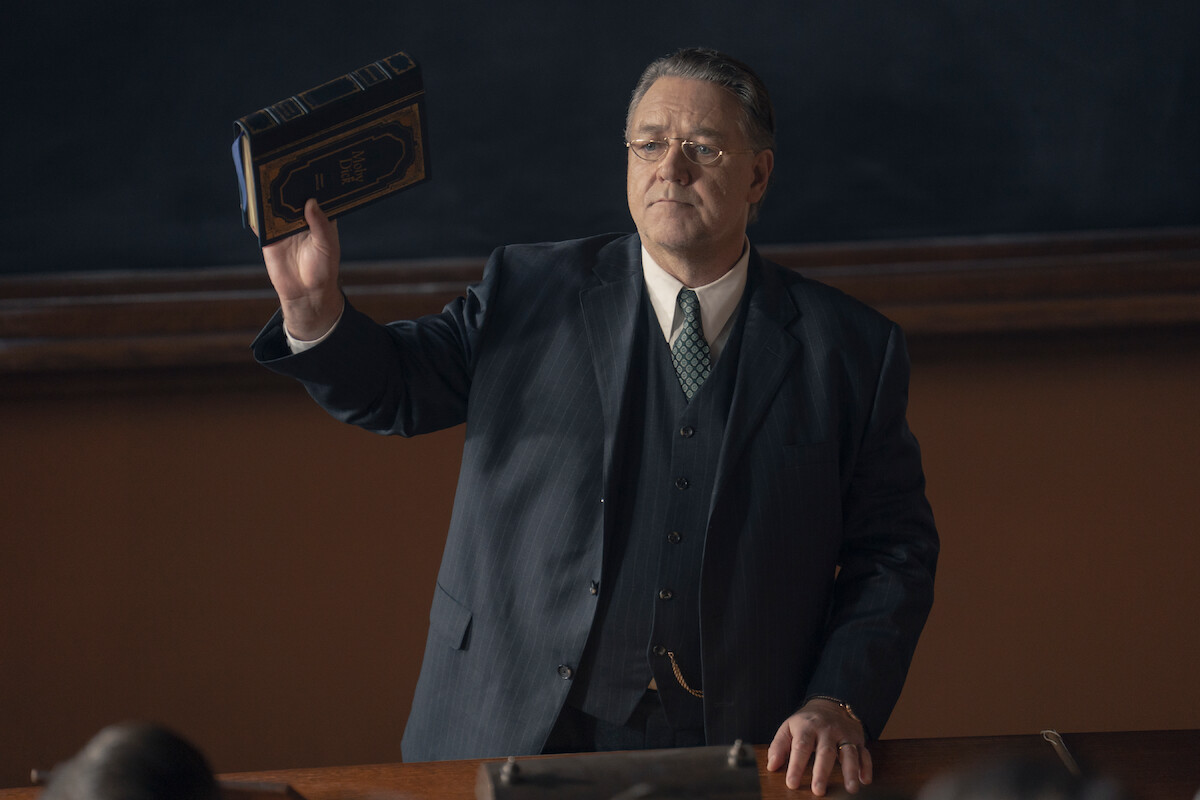 A serious man in a dark suit stands at a desk in a classroom, holding up a book in one hand. He faces the audience, with a blackboard and wood accents in the background, suggesting a formal academic environment.