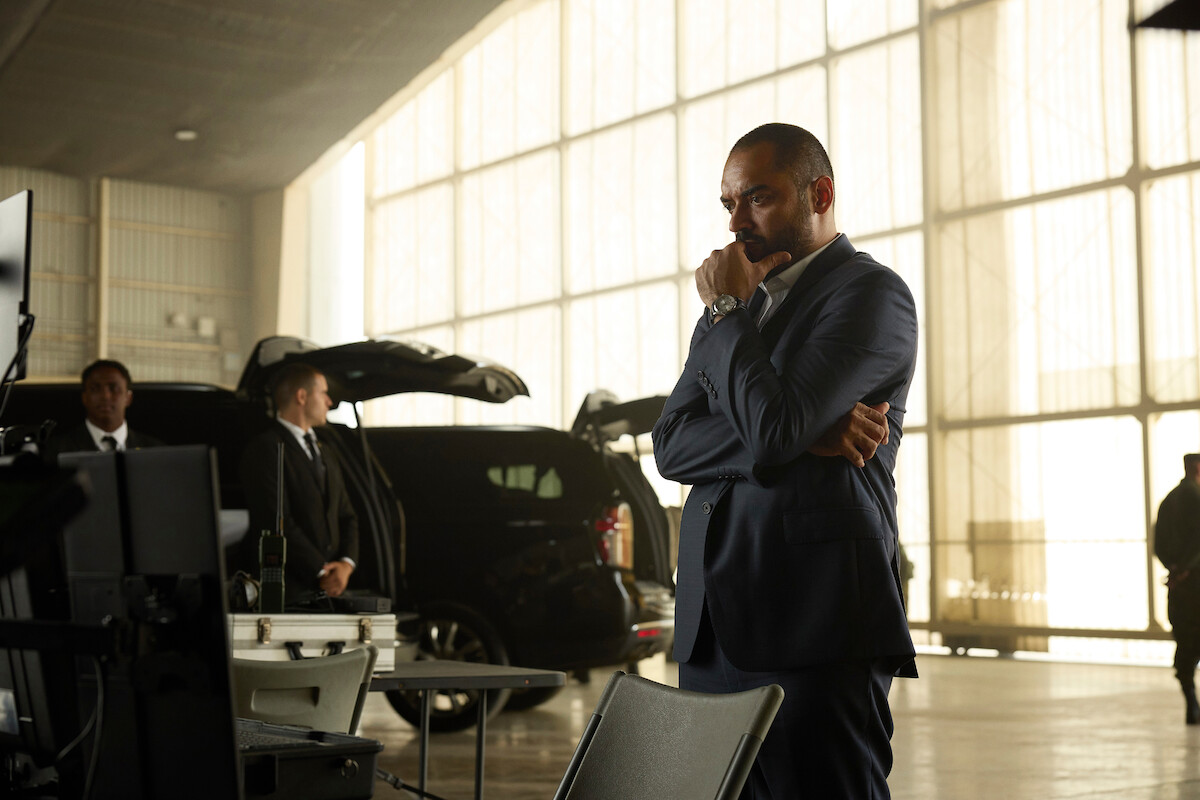 Man in suit standing thoughtfully in a large, sunlit industrial space with security personnel, black SUVs, and open equipment cases, suggesting a high-security or investigative environment.