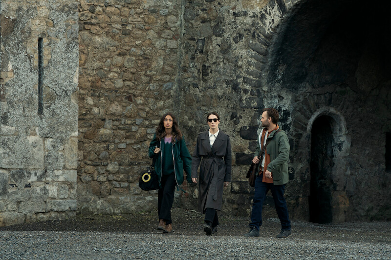 Robyn Cara as Emmy Sizergh, Siobhán Cullen as Dove, and Will Forte as Gilbert Power walk by a large stone wall with arches in 'Bodkin'