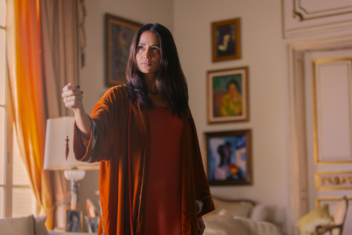 Uma mulher jovem de cabelos longos veste um sobretudo laranja e segura um pêndulo em sua mão, enquanto observa a sala